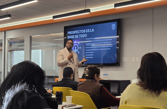 A presenter in a beige coat and glasses stands in front of a large digital screen displaying a slide titled 'PROSPECTOS ES LA BASE DE TODO' with bullet points in Spanish about prospecting strategies, including references to social circles, call centers, and social networks, while addressing an audience seated in yellow chairs in a modern office training room with glass walls and overhead lighting.